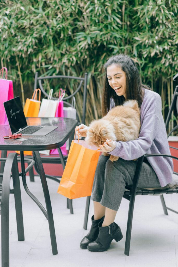 Young woman happily shops online with her pet cat outdoors by a table full of bags.